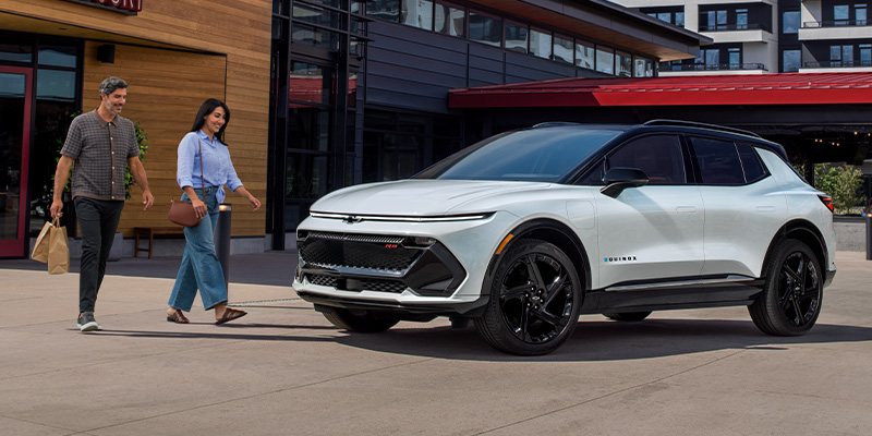 A man and woman walking toward a white Chevrolet Equinox EV parked in a modern outdoor shopping area.