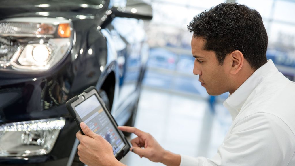 A service technician using a tablet to run diagnostics on a car in a bright dealership service bay.