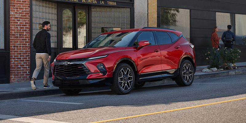 A red Chevrolet Blazer parked on a city street as a man walks past a brick building in the background.