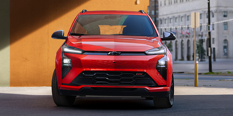 Front view of a vibrant orange-red Chevrolet Bolt EV parked under a shadow on a city street.