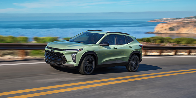 Front-three-quarter view of a sage green Chevrolet Trax driving on a scenic coastal highway alongside the ocean.