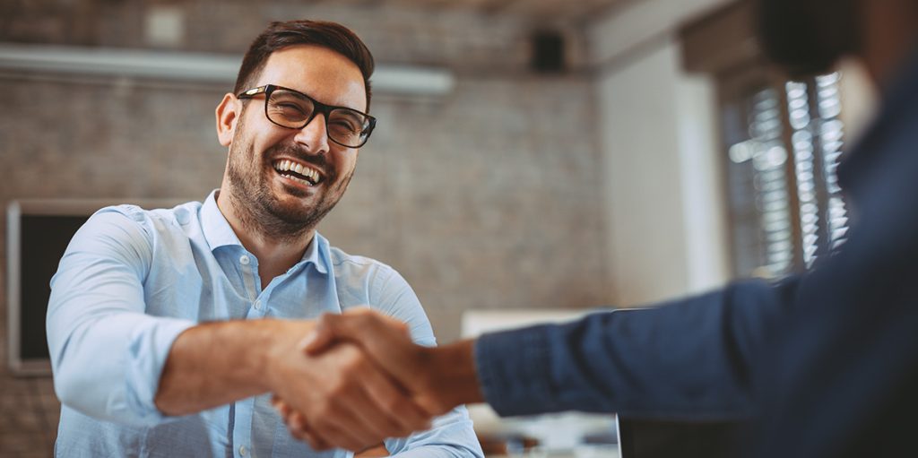 A smiling man with glasses shaking hands with a colleague across a desk in an office setting.