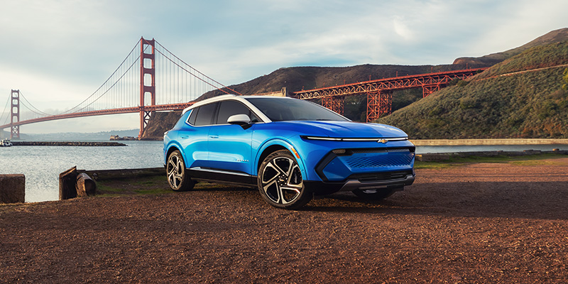 ront-three-quarter view of a bright blue Chevrolet Equinox EV with a white roof parked on a gravel area near the water, with the Golden Gate Bridge visible in the background.